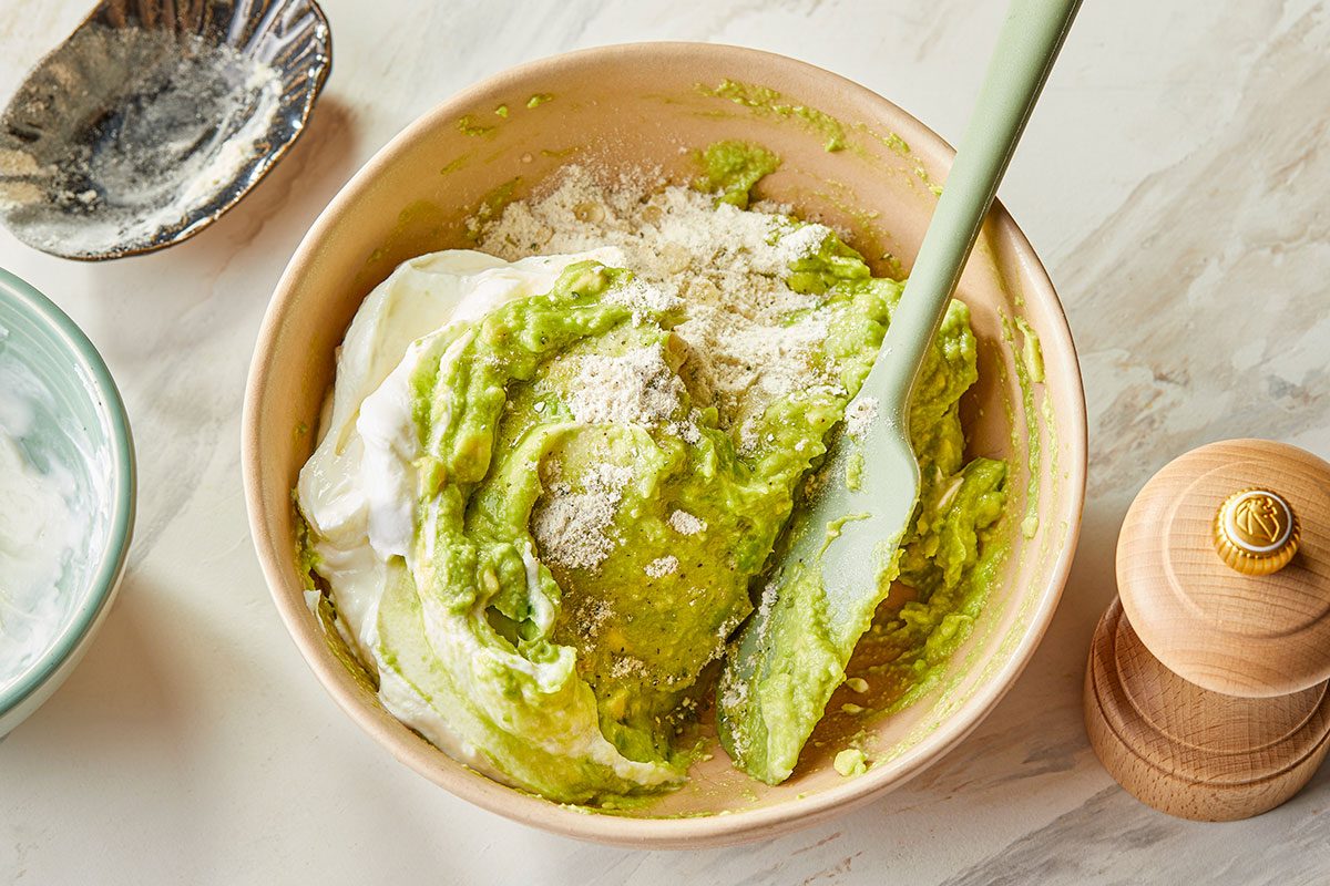 Overhead image showing Greek yogurt and ranch seasoning being added to mashed avocado to create Ranch Avocado Yogurt Dip.