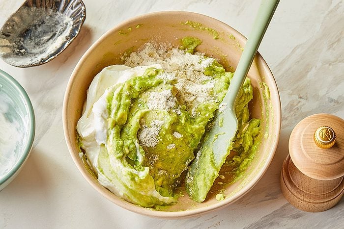 Overhead image showing Greek yogurt and ranch seasoning being added to mashed avocado to create Ranch Avocado Yogurt Dip.
