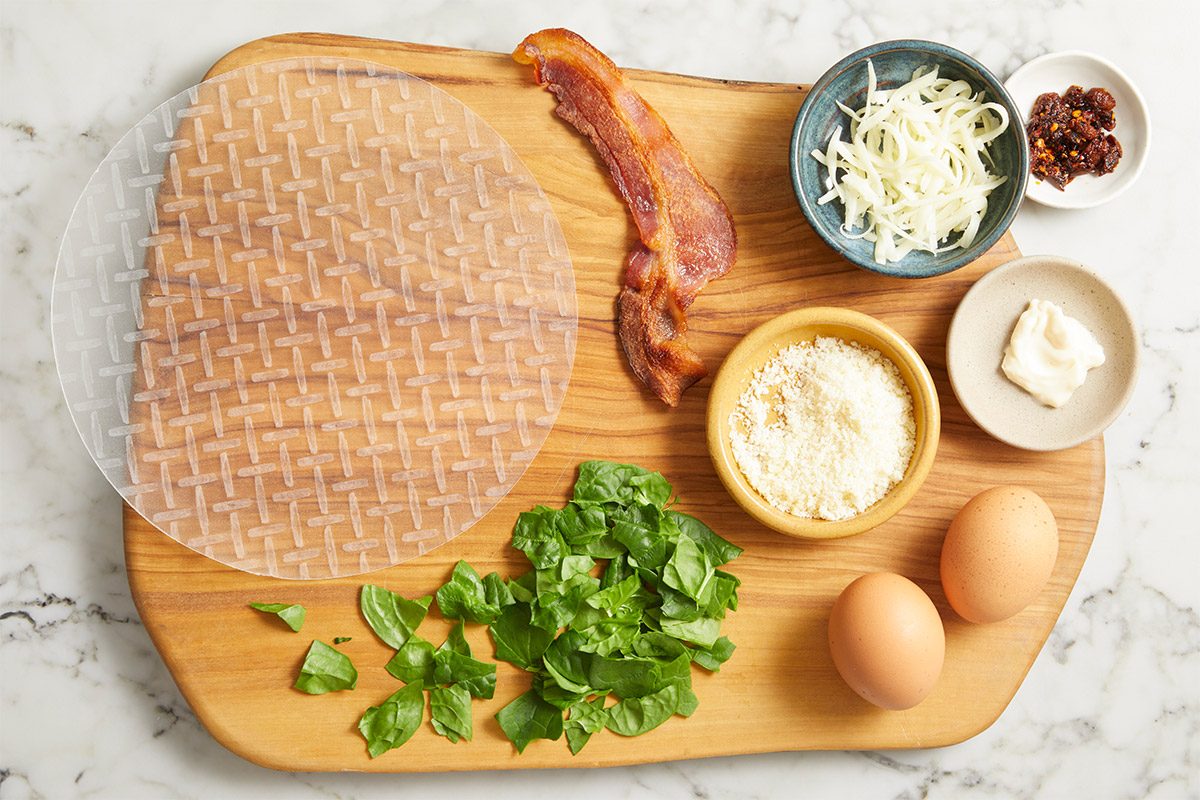 Overhead premium set photography of ingredients arranged on a wooden cutting board for Rice Paper Breakfast Pizza, including eggs, mayonnaise, grated Parmesan, shredded mozzarella, chili crisp sauce, cooked bacon, fresh baby spinach, and an 8-inch rice paper wrapper on a marble surface.