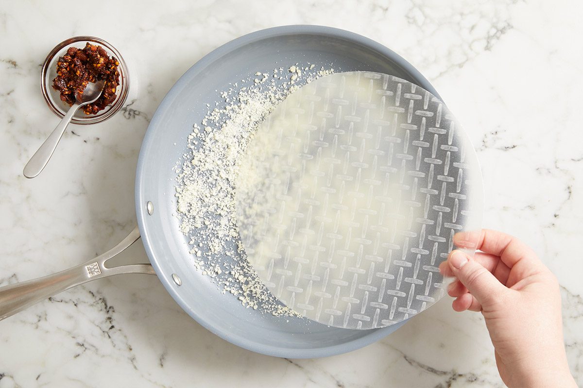 Overhead step-by-step image of rice paper placed into a nonstick skillet as part of preparing Rice Paper Breakfast Pizza, showing the initial stage of cooking on a marble countertop.