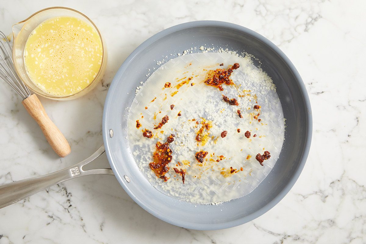 Overhead step-by-step image of rice paper cooking in a skillet topped with grated Parmesan and chili crisp sauce, showing early layering for Rice Paper Breakfast Pizza.