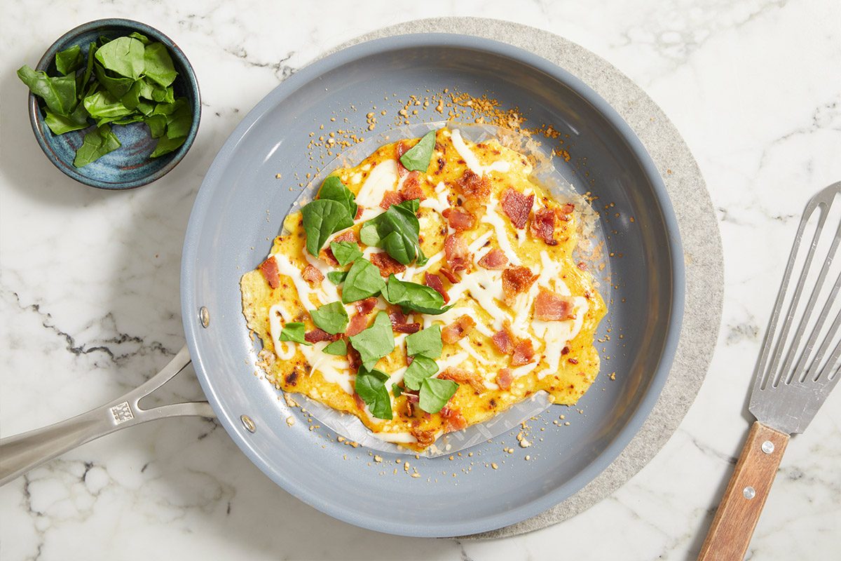 Overhead step-by-step image of Rice Paper Breakfast Pizza cooking in a skillet topped with eggs, bacon, mozzarella, spinach, and chili crisp, nearly finished before folding.