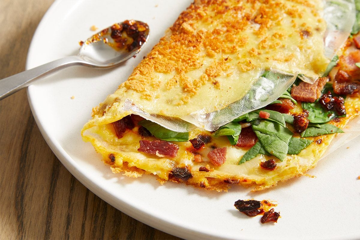 Vertical close-up push and pulls image of Rice Paper Breakfast Pizza on a white plate, highlighting crispy golden rice paper exterior and layered filling of egg, spinach, bacon, and cheese.