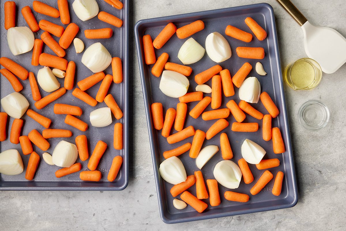 Two baking trays with chopped onions, whole garlic cloves, and baby carrots are arranged on a light countertop. A small bowl of oil and a spatula are nearby, ready for roasting preparation.