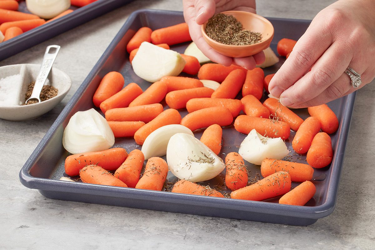 A baking sheet with baby carrots and quartered onions being sprinkled with seasoning by a person’s hands, with bowls of salt and pepper nearby.