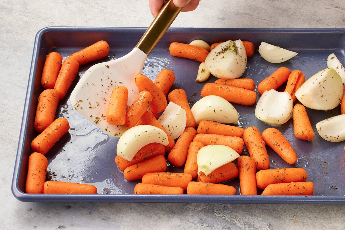 Sliced onions and baby carrots seasoned with herbs on a baking sheet, being mixed with a white spatula.