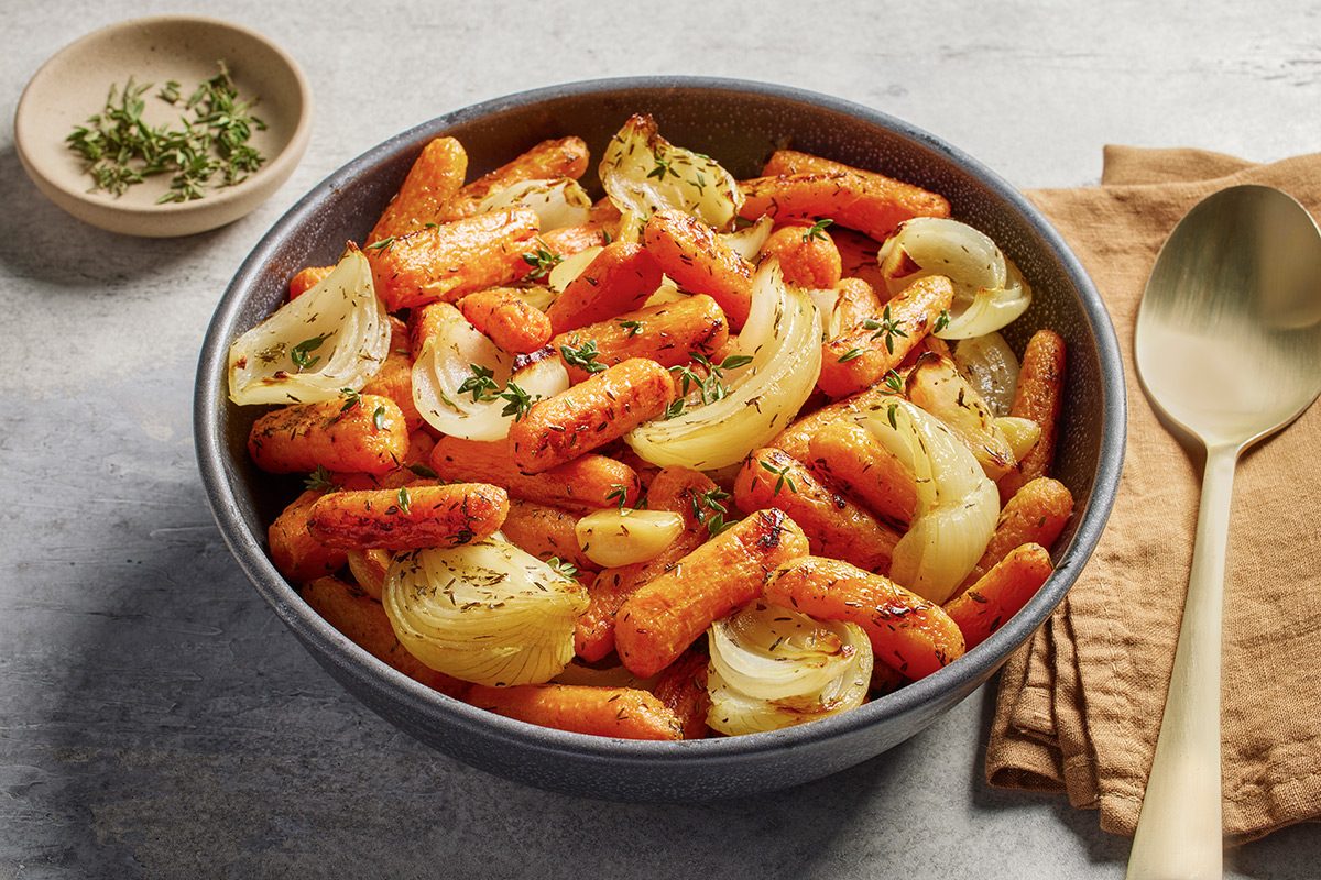 A bowl filled with roasted baby carrots and onion wedges, garnished with fresh herbs. A gold spoon and a small dish of herbs are placed beside the bowl on a brown napkin.