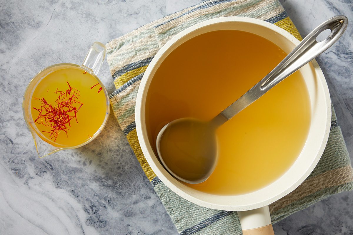 Overhead step-by-step image showing chicken broth infused with saffron threads in a saucepan, creating the golden base used to prepare Saffron Risotto.