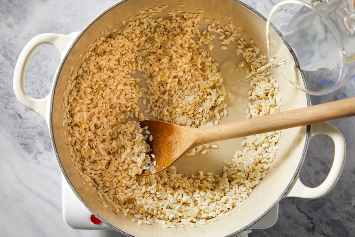 Overhead step-by-step image of arborio rice and finely chopped onion sautéing in olive oil and butter inside a Dutch oven as the risotto cooking process begins.