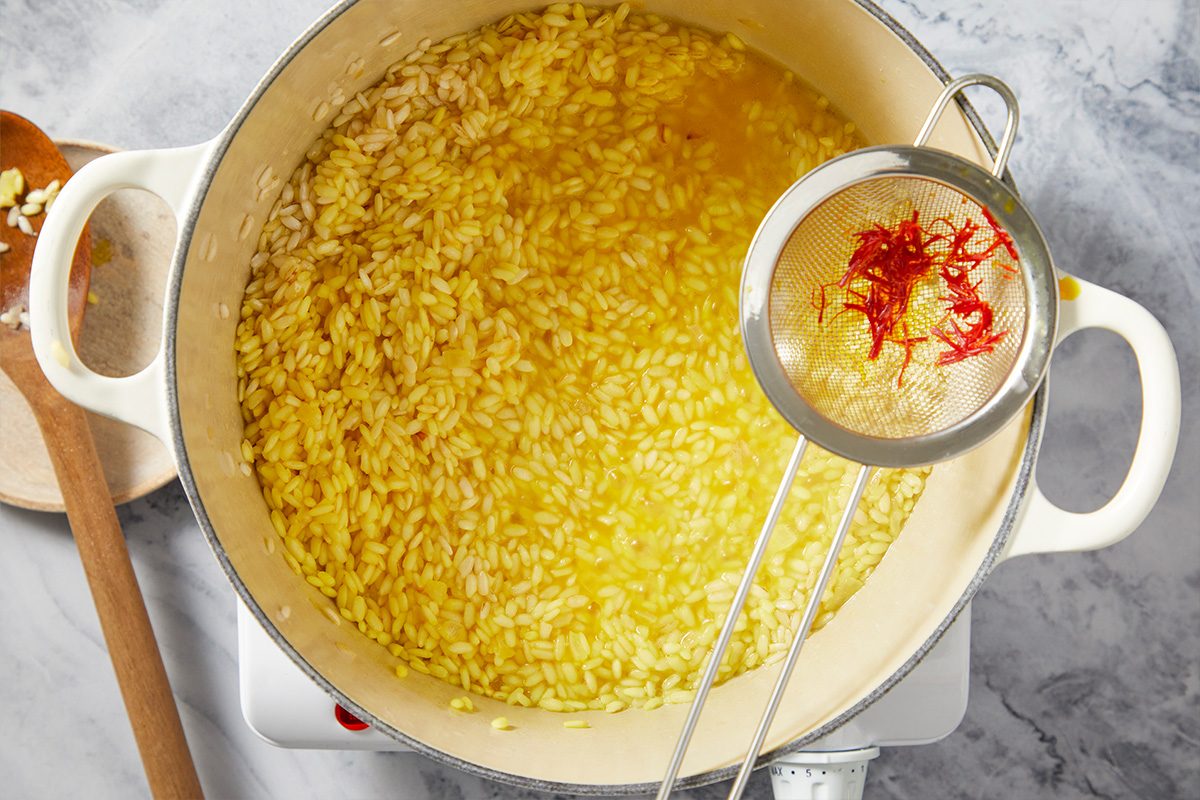 Overhead step-by-step image of saffron broth being poured into arborio rice while cooking risotto, tinting the rice a bright golden color.