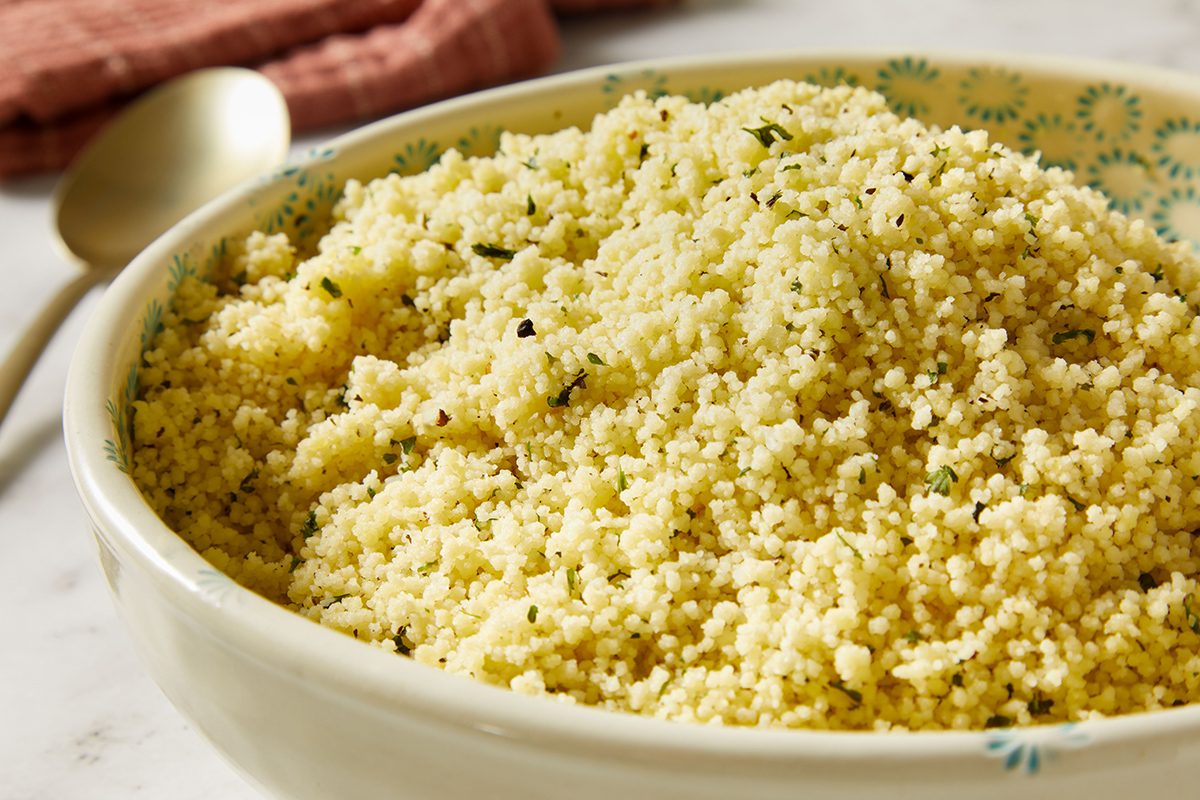 A close-up of a bowl filled with cooked couscous, garnished with herbs, sitting on a light-colored surface next to a spoon and a folded pink cloth.