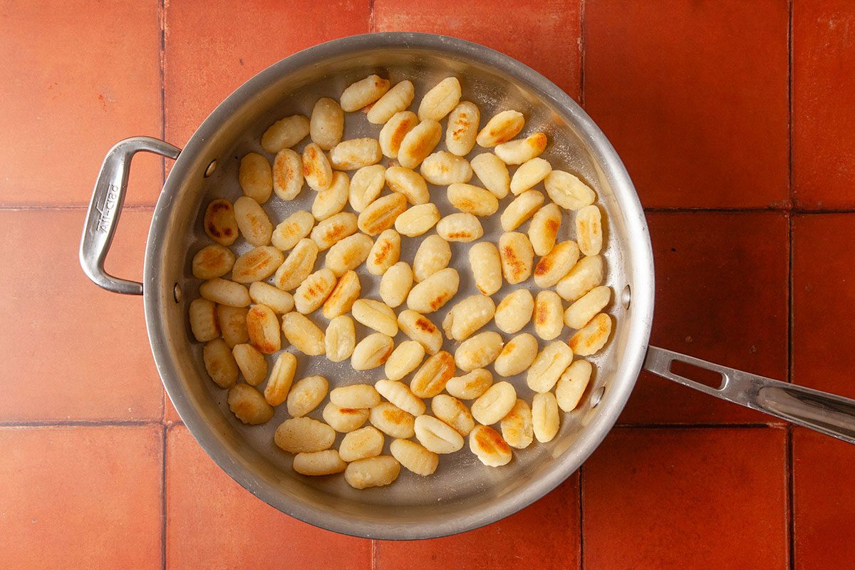 Overhead view of potato gnocchi cooking in a skillet on a terracotta tile surface, showing the start of preparing the gnocchi shepherd’s pie base.
