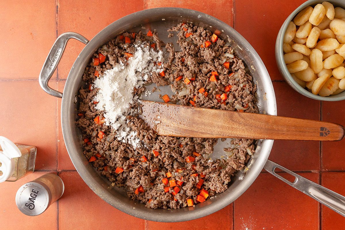 Overhead image of browned ground beef with chopped onions and carrots in a skillet as flour is added to build the savory shepherd’s pie filling.
