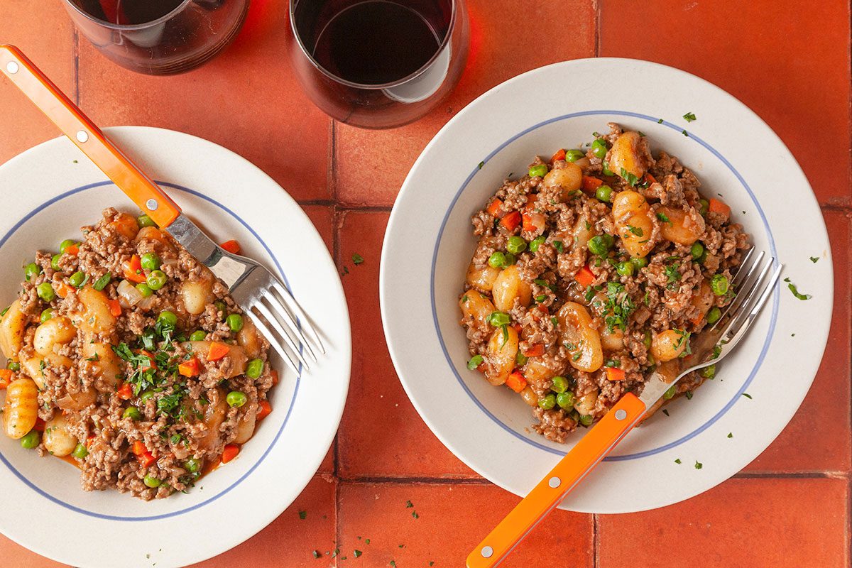 Overhead composition of plated gnocchi shepherd’s pie served in bowls with fresh parsley garnish on a terracotta tile tabletop.