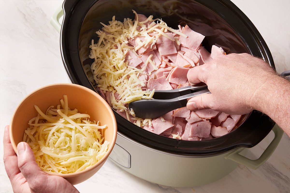 Overhead shot of shredded Swiss cheese being sprinkled over ham and chicken in the slow cooker, with a small bowl of cheese held above the pot.