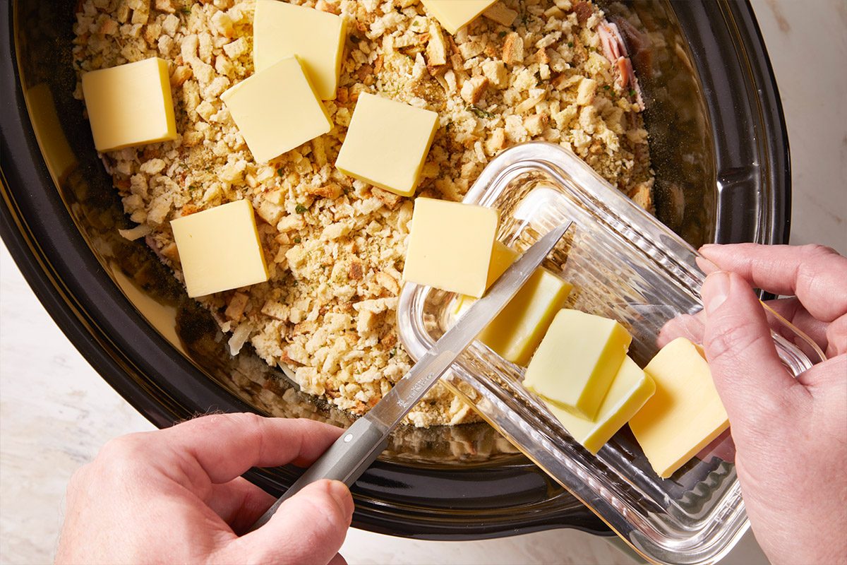 Overhead shot of cubed butter being placed evenly across a chicken mixture in the slow cooker, preparing the dish for slow cooking.