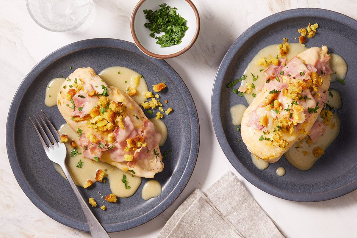 Overhead shot of two plates of Chicken Cordon Bleu arranged side by side, each topped with ham, cheese with a small bowl of chopped herbs in the background.