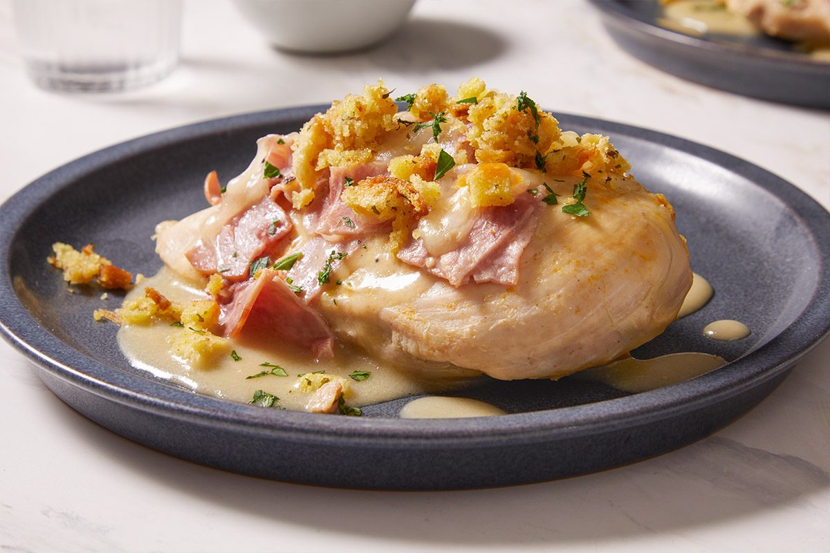 Close-up angled shot of a single serving of Chicken Cordon Bleu on a dark plate, highlighting the juicy chicken, melted cheese, ham slices, and crisp breadcrumb topping