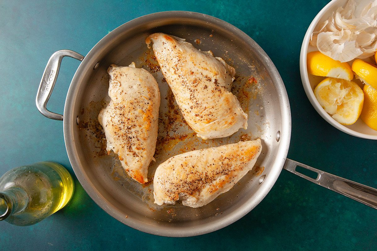 Overhead horizontal image of seasoned chicken breasts searing in a skillet with oil and lemon nearby.