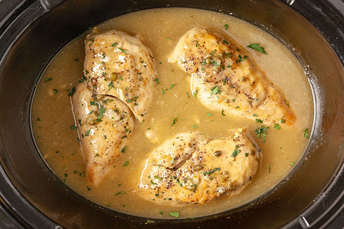 Overhead horizontal image of cooked chicken breasts in slow cooker with thickened lemon caper sauce and parsley garnish.
