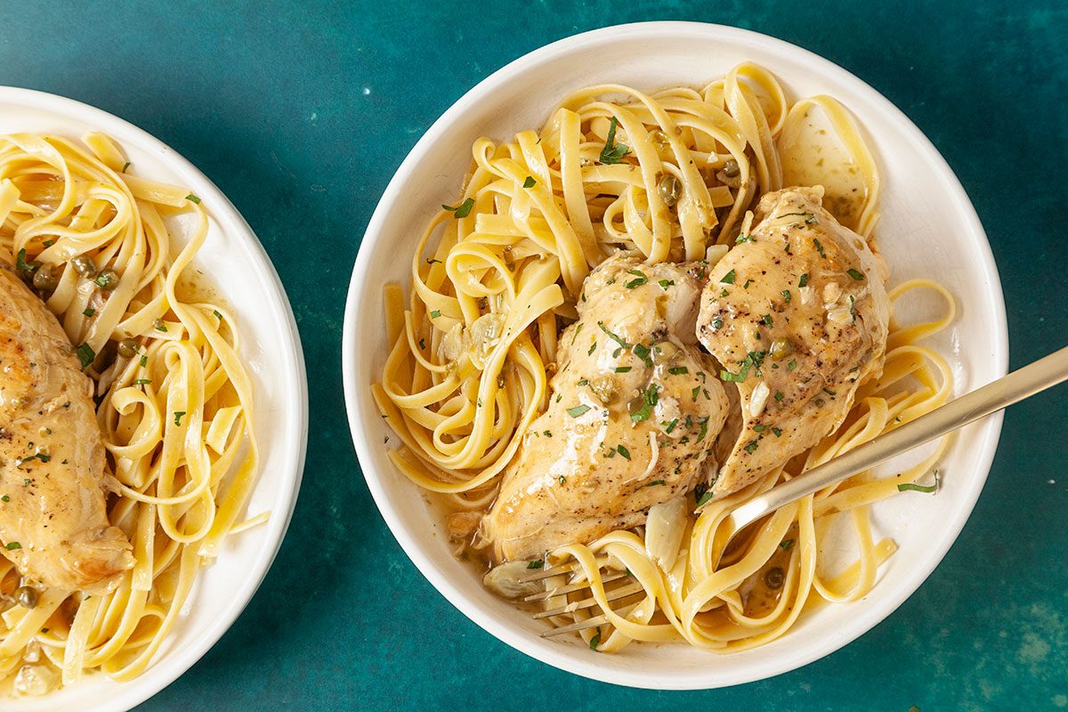 Eye-level horizontal image of chicken piccata served over pasta with lemon caper sauce.