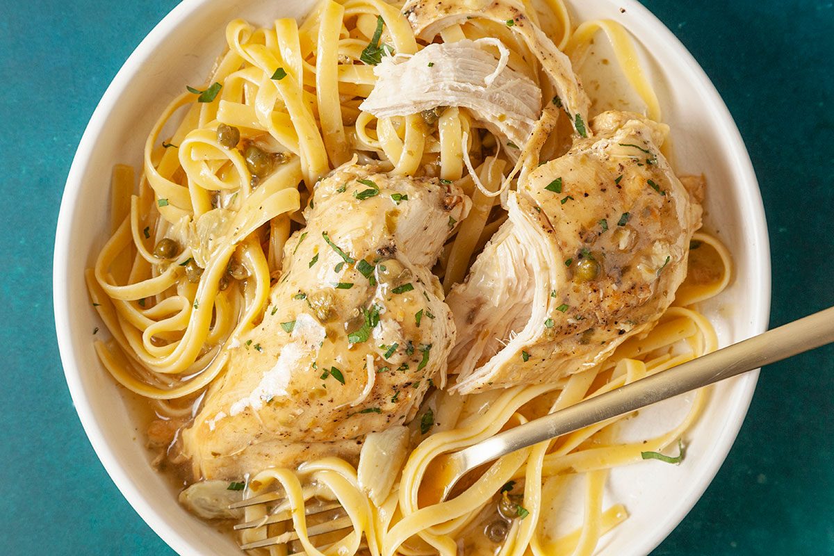 Eye-level horizontal close-up of chicken piccata over pasta showing texture of sauce and garnish.