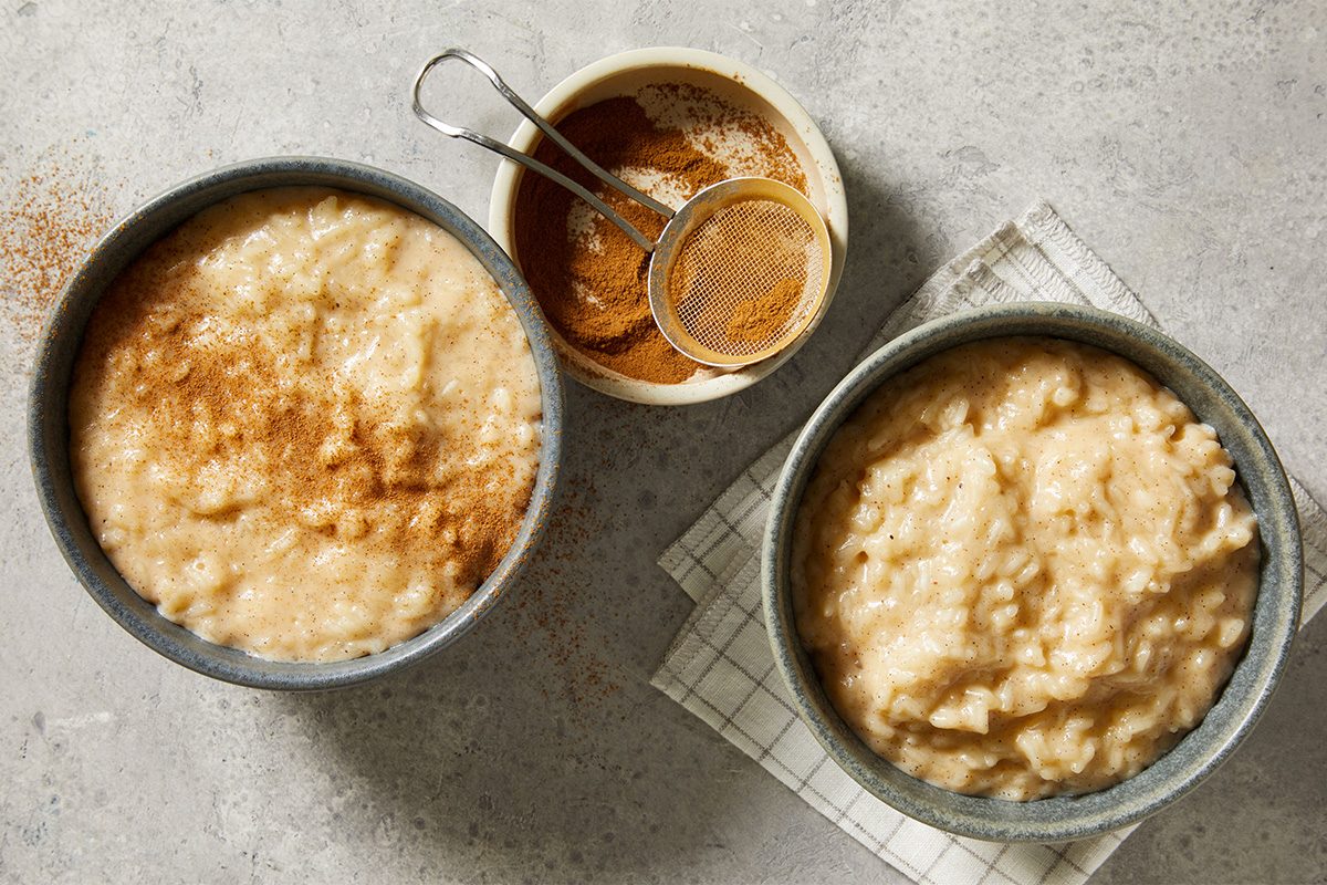 Overhead view of two bowls of Rice Pudding showing a thick, creamy consistency, arranged with cinnamon and spices on the side.