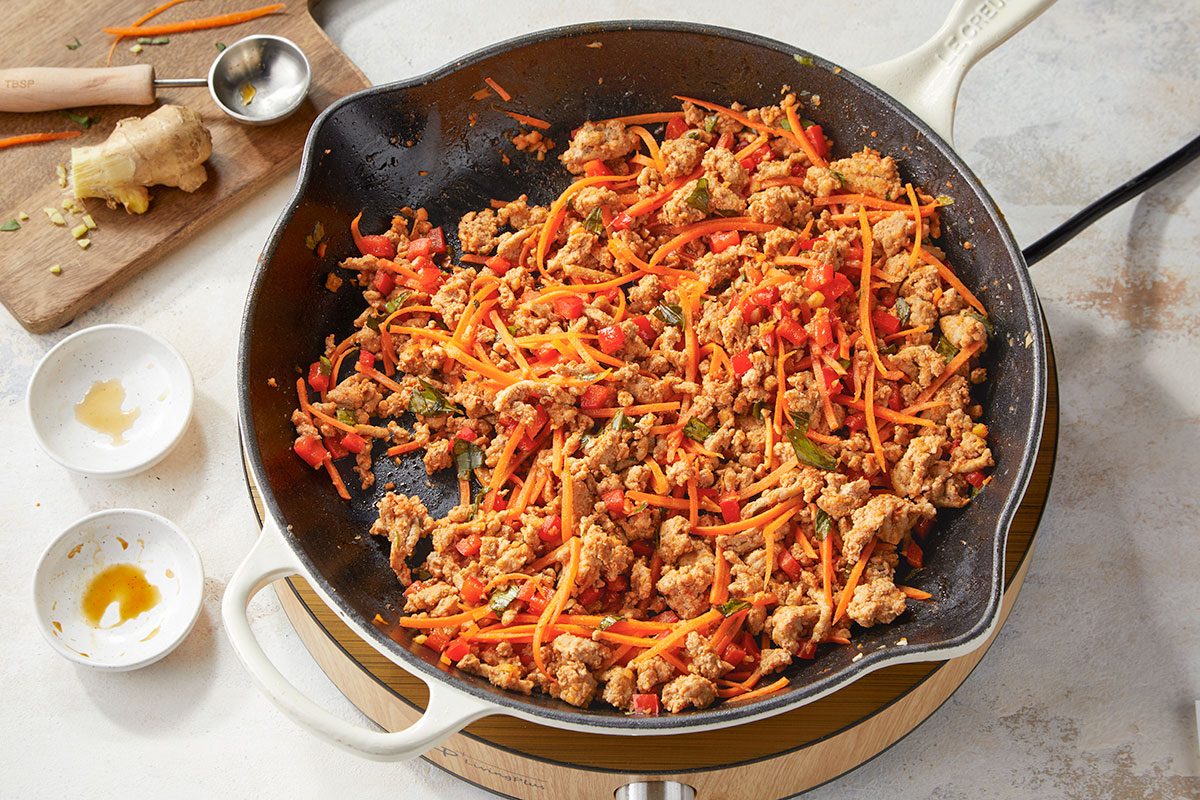 Overhead horizontal image of ground turkey cooking in a skillet with shredded carrots, red pepper, garlic, and ginger.. In a large skillet, cook turkey and red pepper in sesame oil over medium heat until meat is no longer pink. Add garlic and ginger; cook 1 minute longer. Stir in red curry paste, soy sauce, fish sauce, shredded carrots and basil. Heat through