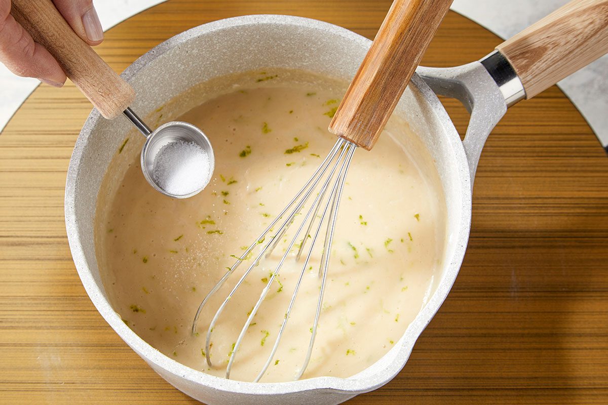 Overhead horizontal image of coconut-peanut béchamel sauce being whisked together in a saucepan.. In a small saucepan, heat oil over medium. Stir in flour and whisk for 1 minute. Slowly add coconut milk, whisking constantly until smooth. Add peanut butter and lime zest. Simmer until slightly thickened, about 3-4 minutes. Season with salt to taste.