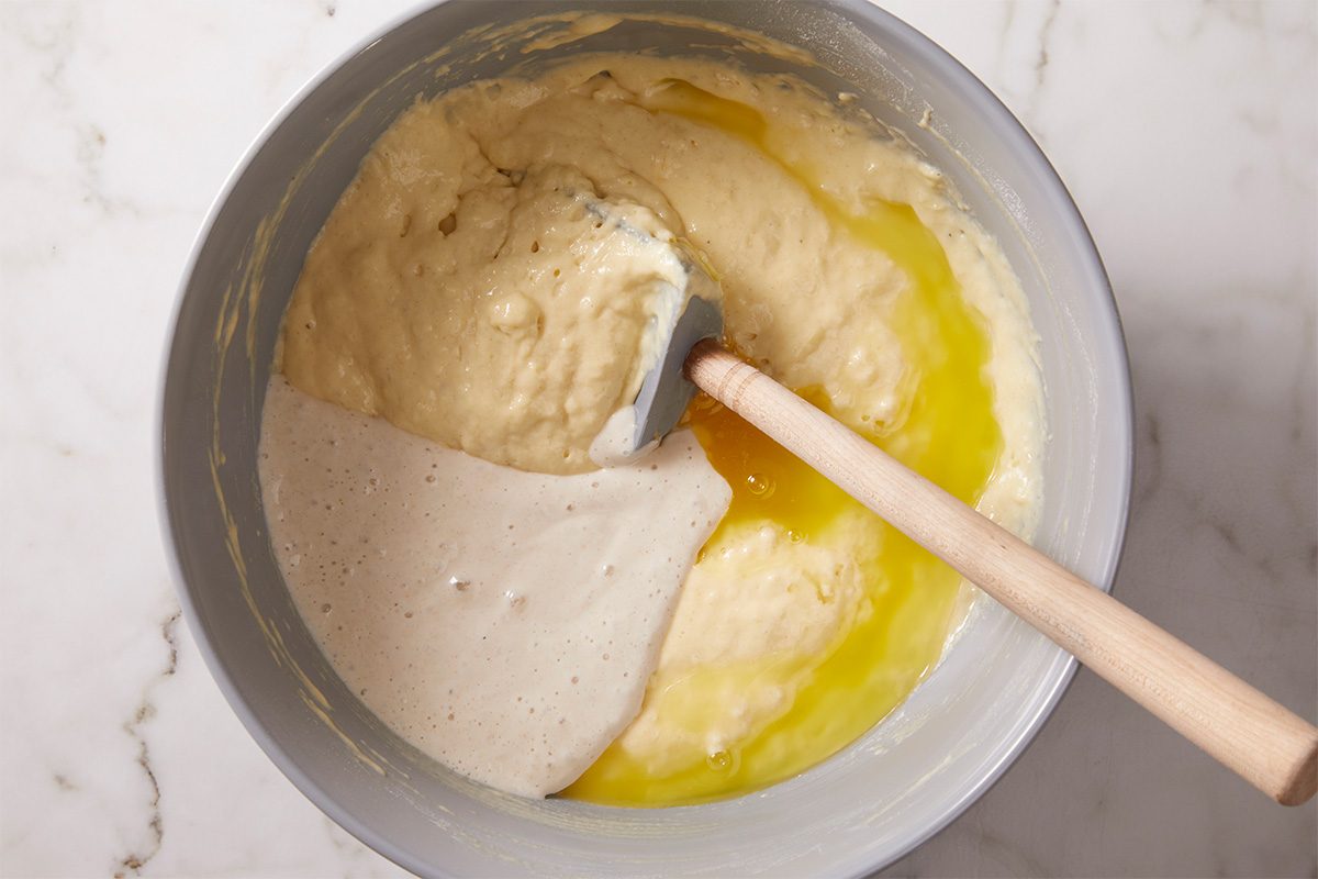 Overhead, horizontal step-by-step image of sourdough waffle batter partially mixed in a bowl. A wooden spoon stirs thick batter alongside melted butter and sourdough starter, showing texture and consistency before cooking.