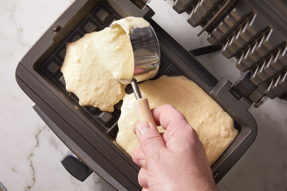 Overhead, horizontal step-by-step image showing sourdough waffle batter being spooned into a preheated waffle iron. Thick batter is spread evenly into the waffle grid before cooking.