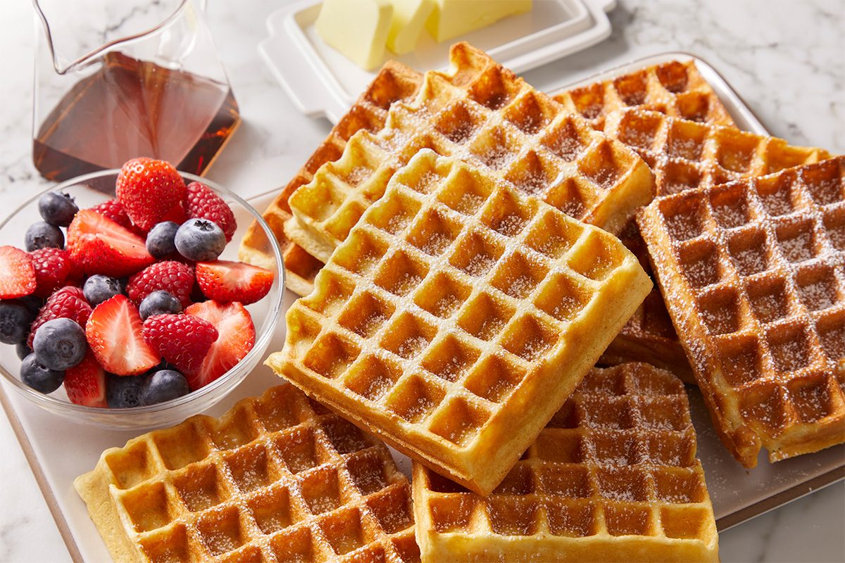 Overhead, horizontal image featuring a stack of golden sourdough waffles served with a glass of maple syrup and fresh berries on a marble surface. Waffles display crisp edges and fluffy interiors.