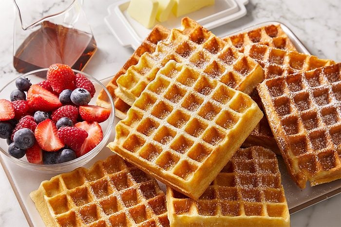Overhead, horizontal image featuring a stack of golden sourdough waffles served with a glass of maple syrup and fresh berries on a marble surface. Waffles display crisp edges and fluffy interiors.