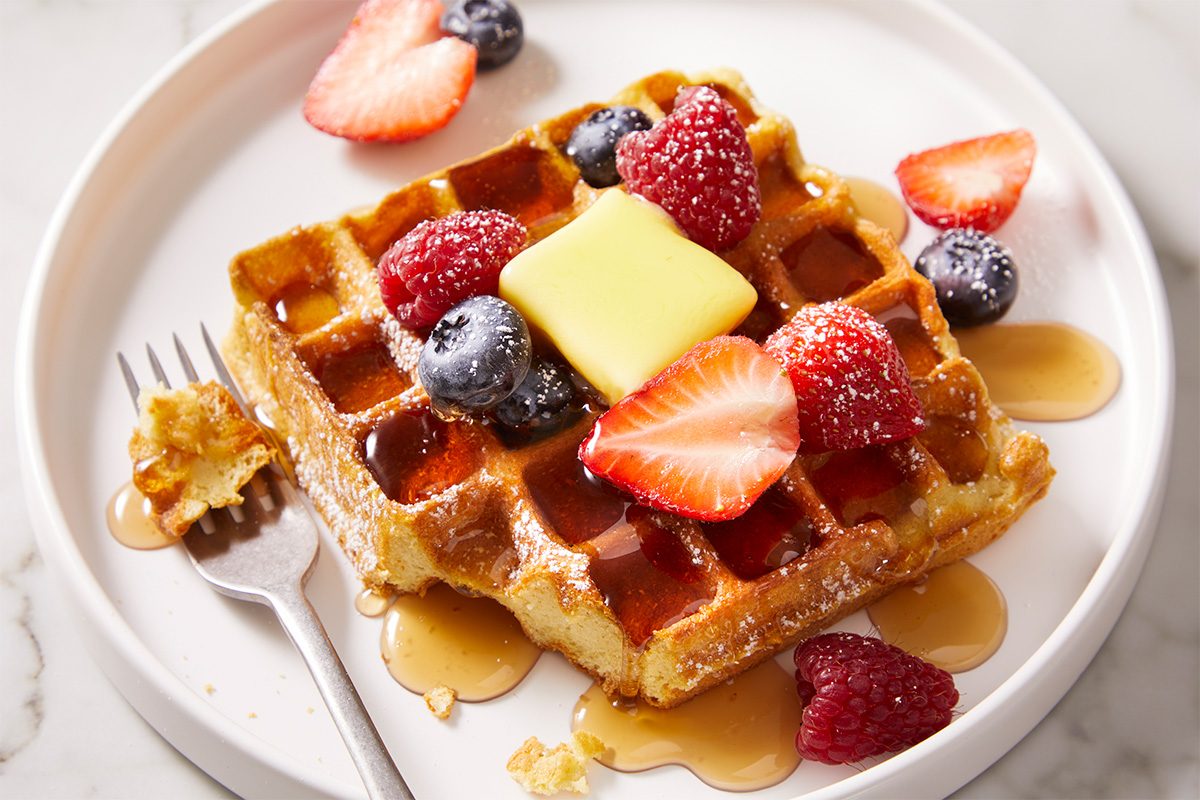 Overhead, horizontal image of plated sourdough waffles topped with butter, fresh berries, and maple syrup. A fork rests on the plate, highlighting a ready-to-eat breakfast presentation.