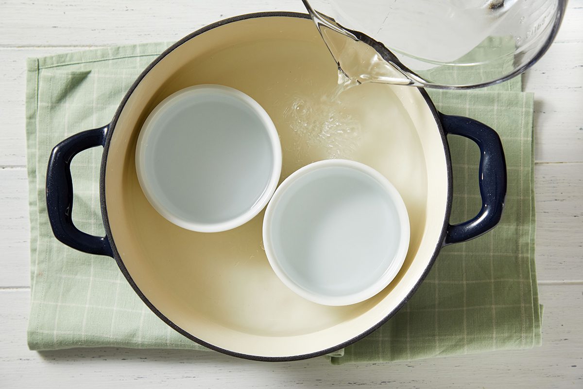 A large cream-colored pot with dark handles contains two small white bowls. Water is being poured into the pot, which sits on a green cloth on a white wooden surface.