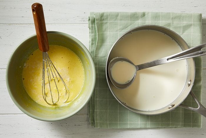 A mixing bowl with a whisk and yellow batter sits next to a saucepan of milk with a metal ladle, both on a green checkered cloth on a white surface.