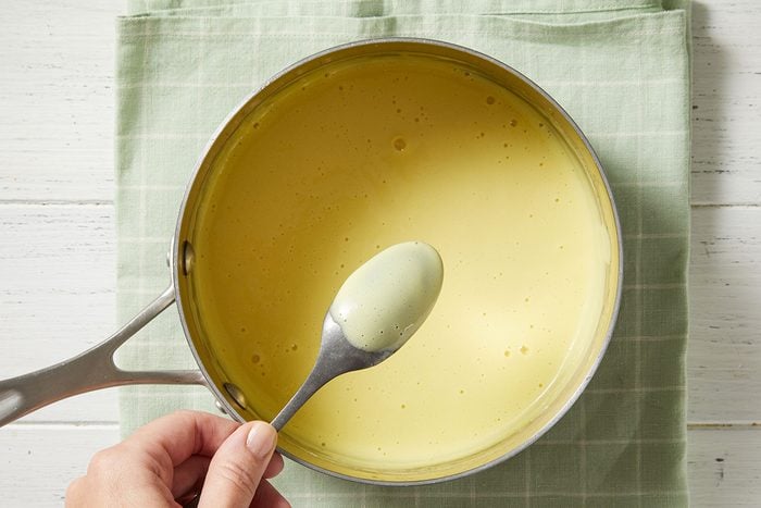 A hand holds a spoon over a saucepan filled with creamy yellow sauce, resting on a light green checked cloth atop a white wooden surface.