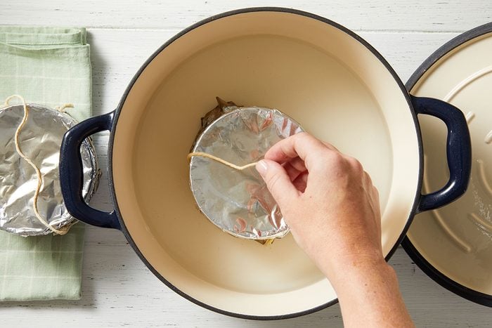A hand places a foil-covered dish with string into a large, empty cream-colored pot with black handles, next to a green cloth on a white surface.