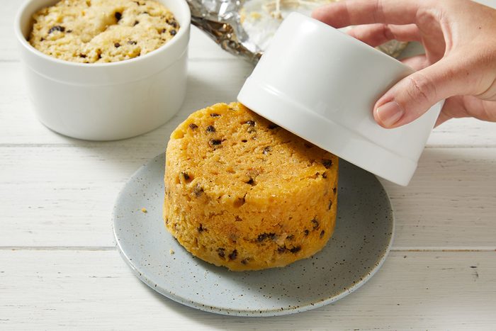 A hand lifts a white ramekin, revealing a round, golden mug cake with chocolate chips on a speckled gray plate. Another ramekin with cake sits in the background on a white wooden surface.