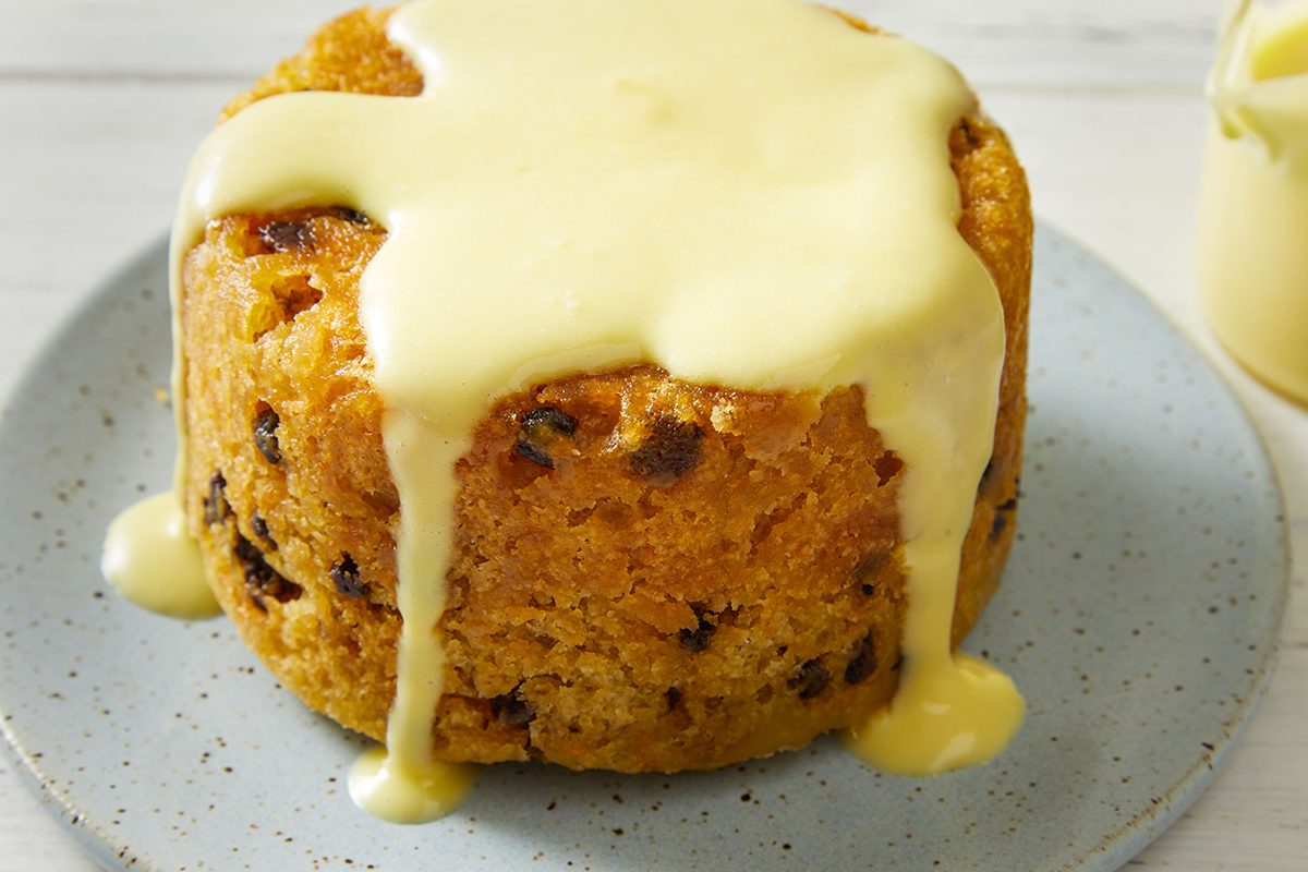 A round spotted dick pudding with visible currants, topped with creamy custard that drips down the sides, served on a light blue speckled plate.
