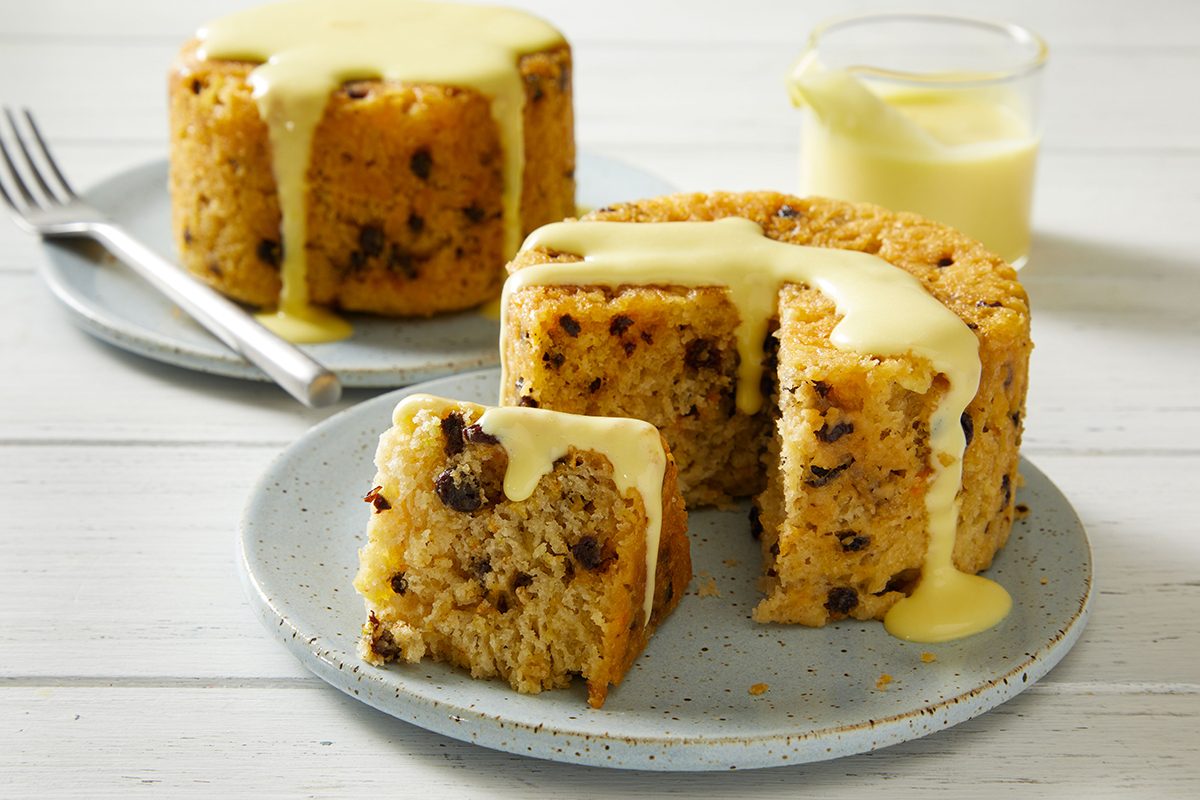 Two round spotted dick puddings with raisins, one partly sliced, are served on plates. Both are topped with creamy custard sauce, with a glass of extra custard in the background on a white wooden table.