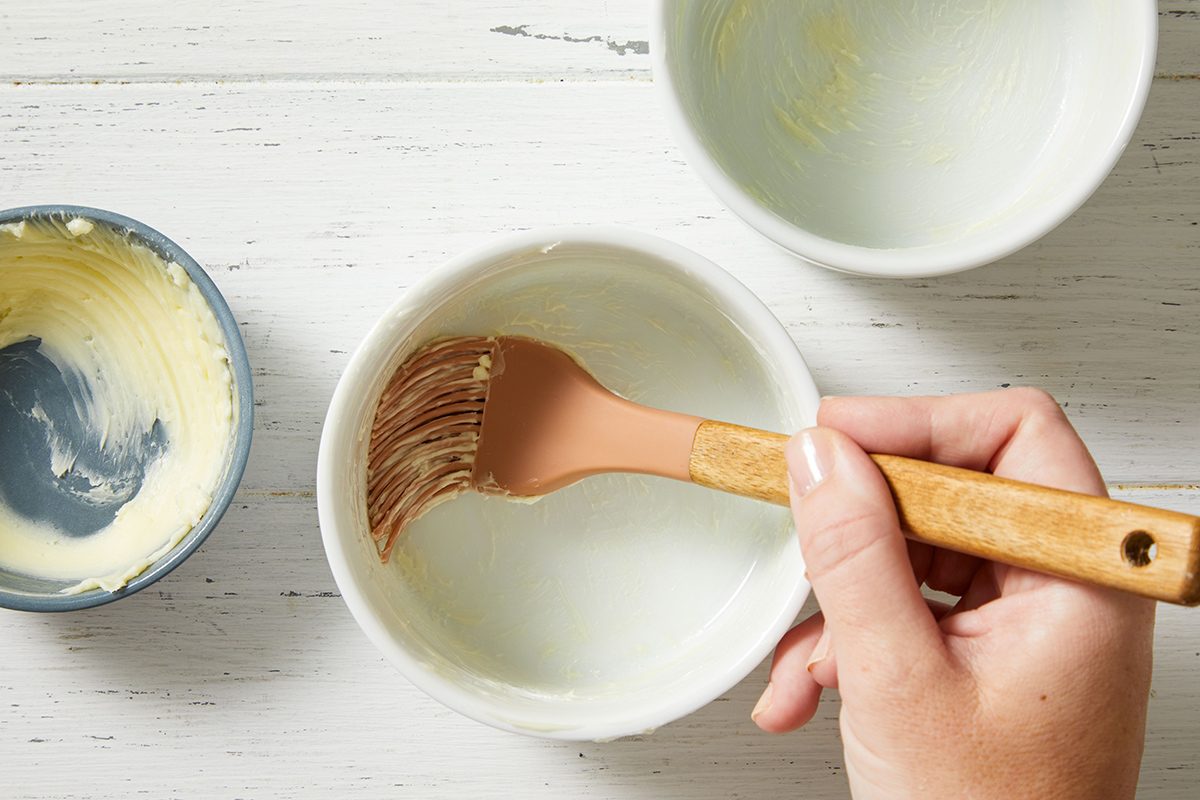 A hand holds a small bowl and uses a silicone brush to spread butter inside it. Two other bowls, also buttered, are nearby on a white wooden surface.