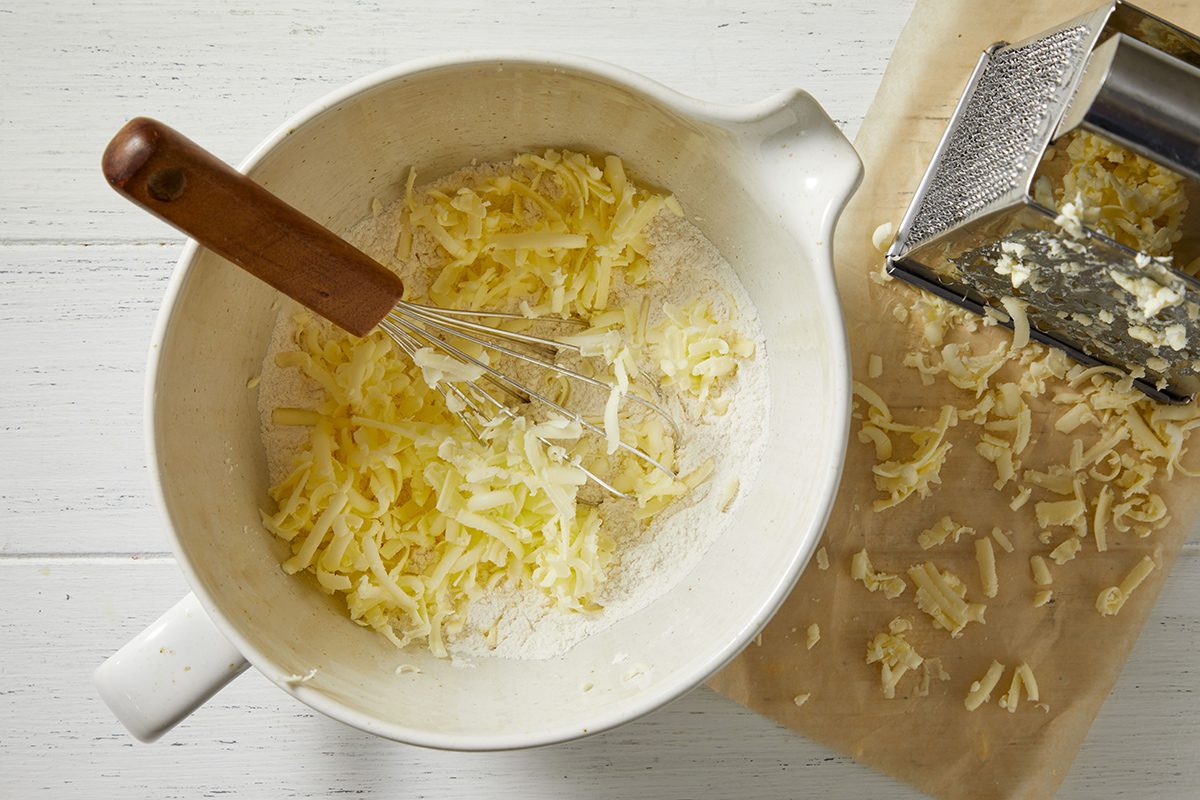 A white mixing bowl with grated cheese and a whisk inside, next to a metal cheese grater and some grated cheese scattered on parchment paper atop a white wooden surface.