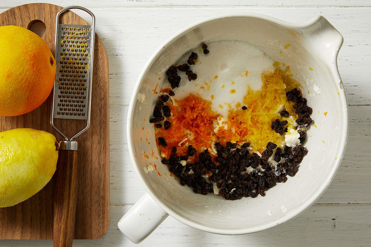 A white bowl containing sugar, currants, and orange and lemon zest sits on a white surface next to a cutting board with whole lemons and a metal grater.