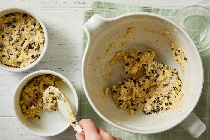 A hand scoops chocolate chip cookie dough from a large mixing bowl into two small white ramekins on a light green cloth and white wooden surface.
