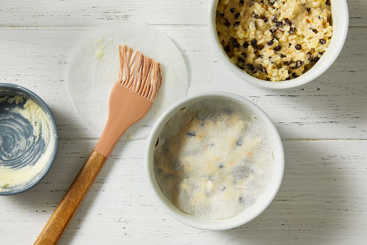 A mixing bowl with dough containing chocolate chips, a pastry brush, a greased ramekin, and a small bowl with remaining butter are arranged on a white wooden surface.