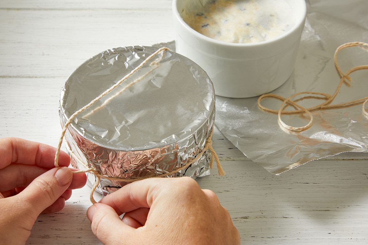 A person ties string around a foil-covered bowl, with a white bowl containing a mixture and some twine on a white wooden surface in the background.