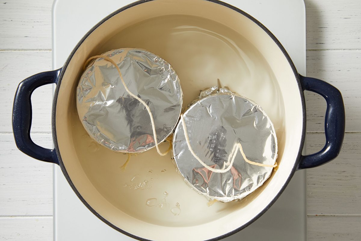 A large white pot with dark handles contains two round, foil-covered dishes tied with string, partially submerged in water, sitting on a white stovetop.