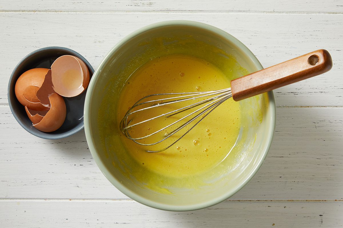 A mixing bowl with a metal whisk and yellow batter sits on a white surface. Next to it is a small bowl containing brown eggshells.