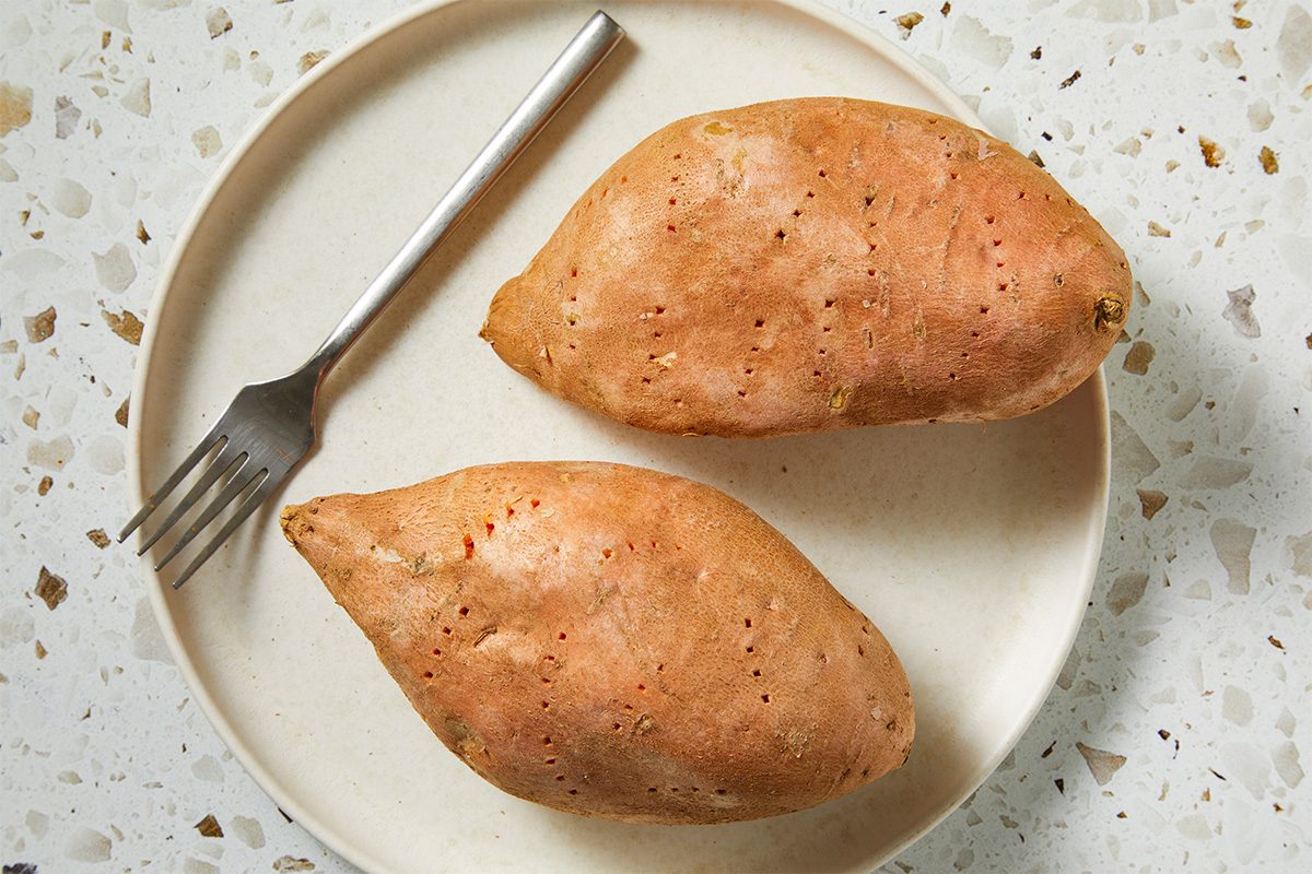 Overhead horizontal image of two whole steamed sweet potatoes resting on a neutral plate with a fork nearby. The simple composition highlights the base ingredient before smashing and roasting, emphasizing texture and natural color in a clean, modern setup.